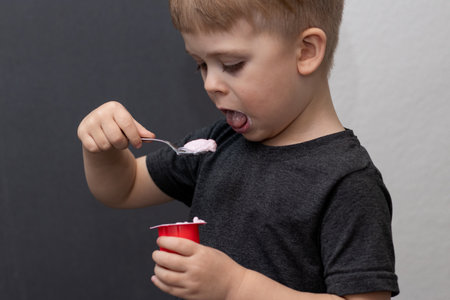 Side portrait of a cute toddler boy eating yoghurt with a spoon, wearing grey shirtの写真素材