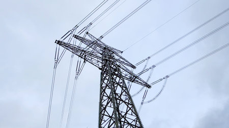 High-voltage electricity transmission tower with multiple power lines against a cloudy sky,...