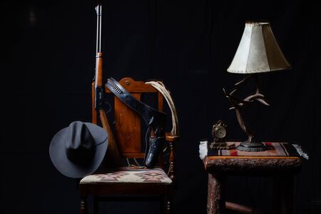 Lever action rifle, revolver, rope, cowboy hat, clock, table, lamp and a chair on black