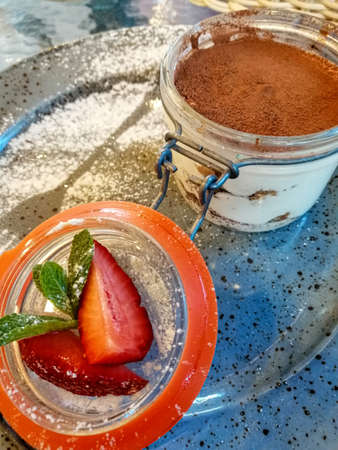 On a blue plate, sprinkled with powdered sugar, tiramisu cake in the shape of a cup and slices of fresh strawberries with mint.の写真素材