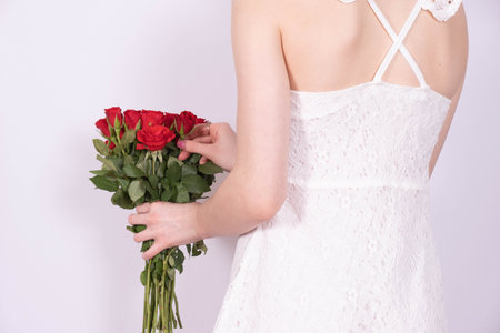 a young girl in a white dress turned her back and holds a bouquet of red roses in her hand on a white backgroundの写真素材