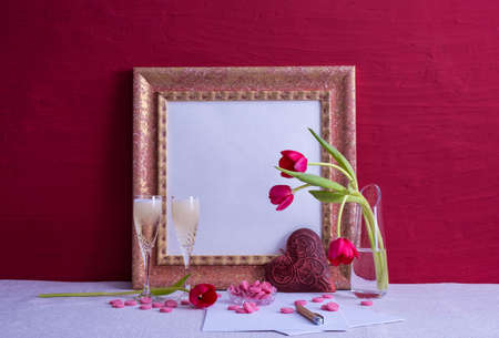 two glasses with champagne, tulips in a vase, an iron heart, an old photo frame on a red background on the tableの写真素材