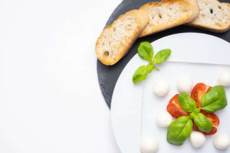 small mozzarella, fresh basil and chopped tomatoes on a white plate and on a stone stand croutons on a white plateの写真素材