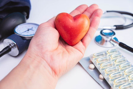red stone heart held in hand, stethoscope, pills and pressure meter on white background on table