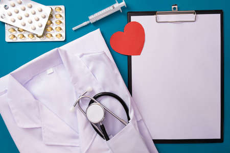 white uniform of a doctor and a stethoscope lie on the table, next to a red heart, pills, on a blue backgroundの写真素材