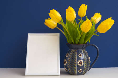 ceramic jug with beautiful yellow tulips and a white frame with a white background, on a blue backgroundの写真素材