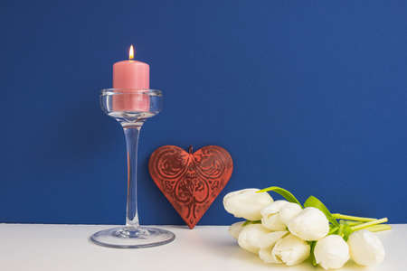 a bouquet of white tulips, a glass candlestick with a burning candle and a decorative metal heart, burgundy on a blue backgroundの写真素材