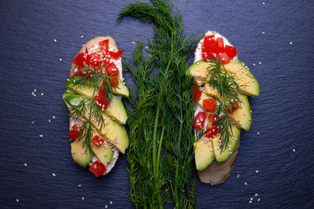 two slices of baguette with slices of fresh avocado and slices of ripe tomato, sprinkled with sesame seeds, canapes and fresh dill on a black backgroundの写真素材
