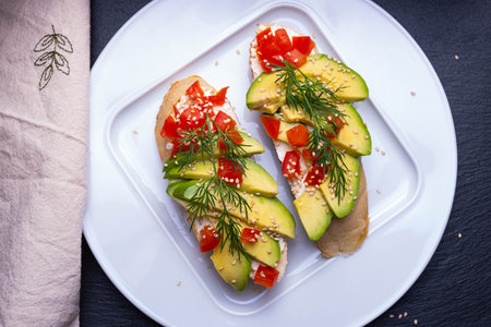 Slices of fresh avocado and slices of ripe tomato are laid out on a slice of baguette and sprinkled with sesame seeds, on a white plate and a napkin, on a black backgroundの写真素材