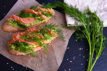two slices of crispy baguette greased with avocado and decorated with smoked salmon fillet and fresh dill sprigs, a napkin and a dill sprig next to it, black background and close-upの写真素材