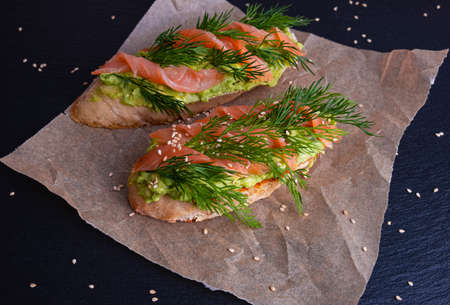 two pieces of crispy baguette greased with avocado and decorated with smoked salmon fillets and sprigs of fresh dill, lie on parchment paper, black backgroundの写真素材