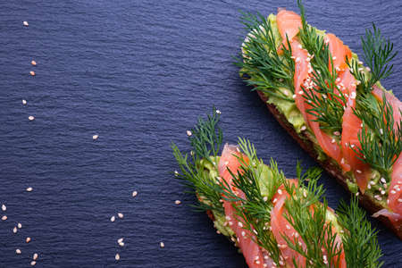 two mouth-watering crispy avocado baguette slices with smoked salmon fillet and fresh dill sprigs and sprinkled with sesame seeds, close-upの写真素材