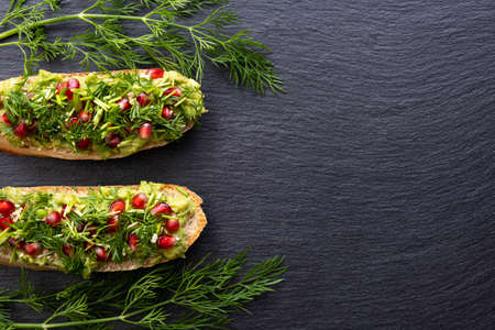two snacks - avocado, sprinkled with ripe pomegranate seeds, sesame seeds and fragrant fresh dill, lie on a black background and dill sprigs on the sides, close-upの写真素材