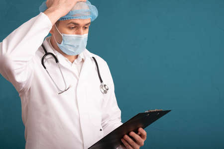 doctor in a white coat and a stethoscope on his shoulders holding a folder and holding his head,...