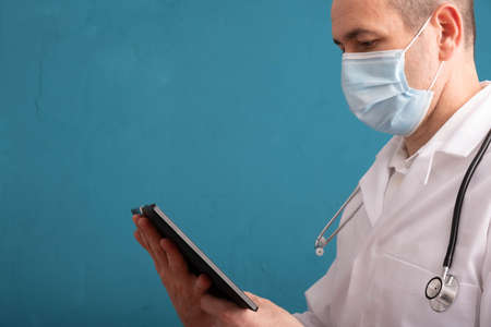 a doctor in a mask and a stethoscope on his shoulders, looks at the tablet, on the left side of the...