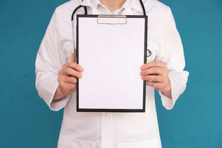 doctor shows folder with blank sheet of paper, stethoscope on shoulders, blue backgroundの写真素材