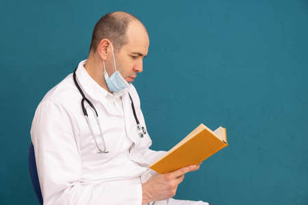doctor in a mask and a stethoscope on his shoulders, reads a book, on a blue backgroundの写真素材