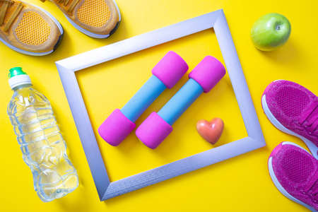metal frame, pink dumbbells and heart, green apple and bottle of water, yellow sneakers and yellow backgroundの写真素材