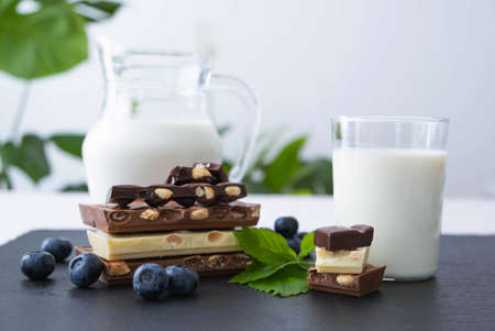 broken bars of black, white and milk chocolate with blueberry berries nuts and a jug of milk on a...