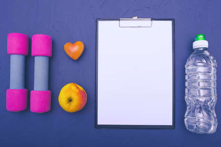 sports equipment - dumbbells, an apple and a bottle of water and a blank sheet of paper to record achievements, blue backgroundの写真素材