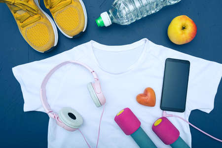 white t-shirt and yellow sneakers, bottle of water and apple, dumbbells, cell phone with headphones, blue backgroundの写真素材