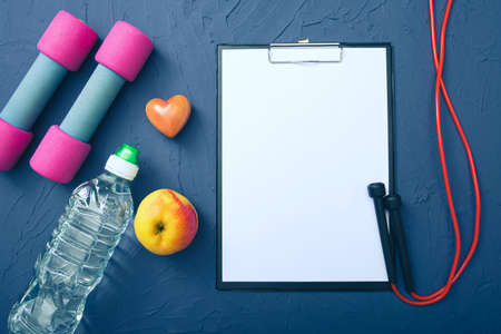 sports equipment - dumbbells, an apple and a bottle of water and a jump rope, a blank sheet of paper to record achievements, blue backgroundの写真素材