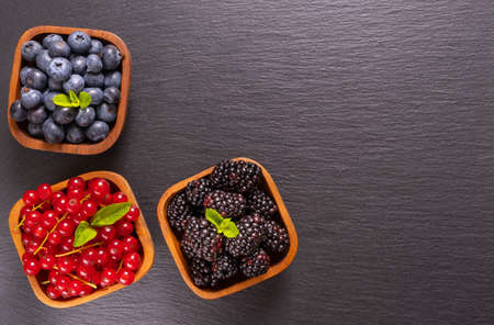 full wooden bowls of ripe berries - blueberries, currants and blackberries and mint leaves on a black backgroundの写真素材