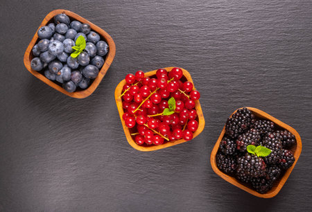 full wooden bowls of ripe berries - blueberries, currants and blackberries and mint leaves on a black backgroundの写真素材