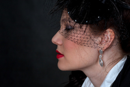 Portrait of the young girl sitting on a chair in a black jacket and a hat with a veil on a black...