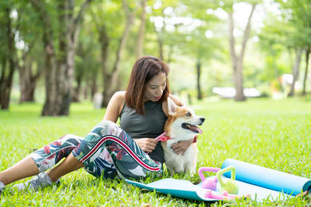 A healthy woman playing with a corgi puppy while excersing on yoga mat surrounding with gym tools such as kettlebell and dumbbell, outdoor training with dogの写真素材