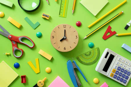 Different stationery and clock on green table background, flat lay. Space for text, back to school...
