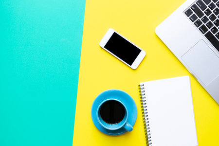 Coffee cup, note book, computer and mobile phone on the yellow and green table in the office backgroundの写真素材