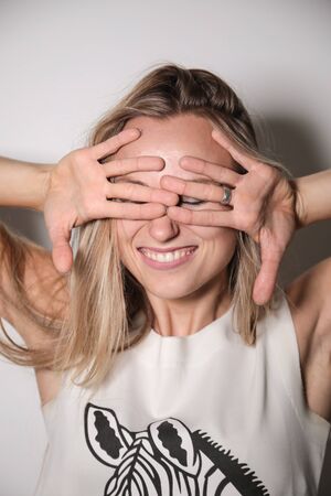 A young model, blonde, dressed in a T-shirt with cut-out sleeves, facing a white wall and illuminated by a strong flash light, smiles as she covers her eyes with both hands outstretched.の写真素材