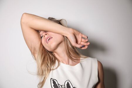 A young model, blonde, dressed in a T-shirt with trimmed sleeves, placed in front of a white wall and illuminated by a strong flash light, covers her face with her right arm.の写真素材