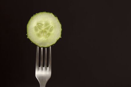 Slice of fresh green cucumber on fork on dark black background. silhouette of a fork.の写真素材