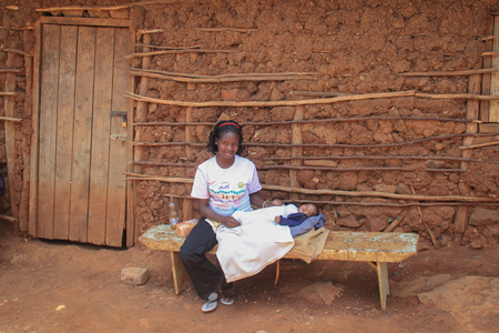 Kibera, Nairobi, Kenya - February 13, 2015: African woman in a white T-shirt holding a baby in her arms and sitting on a bench near a clay hutのeditorial素材