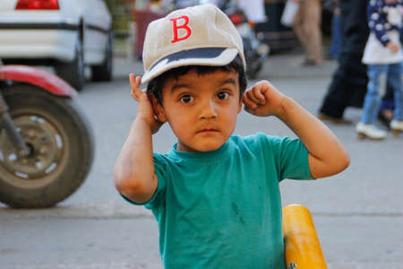 Shiraz, Iran - May 10, 2017: a cute Iranian baby boy in a cap and a bright T-shirt with huge brown eyes smiles shyly and poses for the camera in the city.のeditorial素材