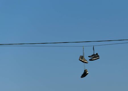 Old ripped sneakers hang on electric wires against the blue sky in the city. Hooliganism.の写真素材