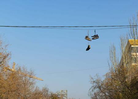 Old ripped sneakers hang on electric wires against the blue sky in the city. Hooliganism.の写真素材