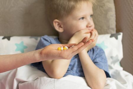 A sick sad child with a temperature and a headache lies in bed, refuses to drink medicine and pills. Flu colds disease virus bacteriumの写真素材