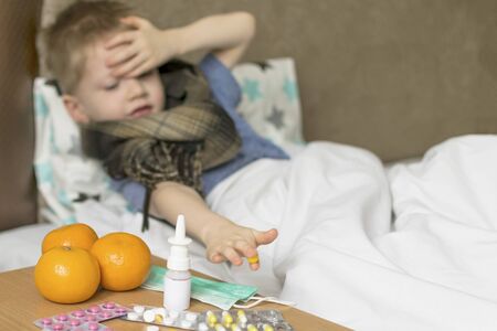 A sick sad child with a temperature and a headache lies in bed next to medication. Flu colds disease virus bacteriumの写真素材