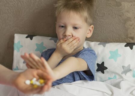 A sick sad child with a temperature and a headache lies in bed, refuses to drink medicine and pills. Flu colds disease virus bacteriumの写真素材