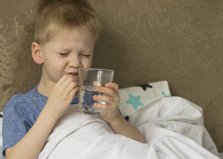 A sick sad child with a temperature and a headache lies in bed next to medicines and pills. Flu colds disease virus bacteriumの写真素材