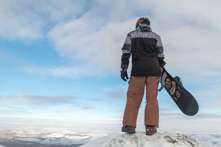 The snowboarder stands on the edge and holds the snowboard. The skier climbed the mountain. Freeride in the mountains. A young man extreme stands with a snowboard on top of a rock.の写真素材