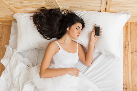 Young attractive woman sleeping in bed alone holding mobilephoneの写真素材