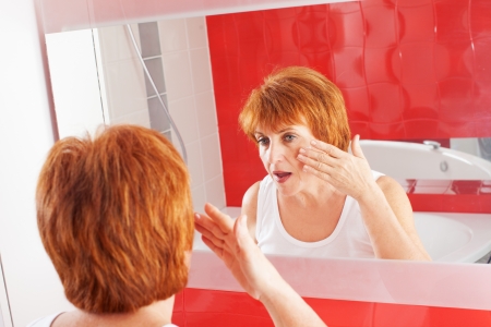 Mature woman gets cream on face in bathroom. Adult female looking at mirrorの写真素材