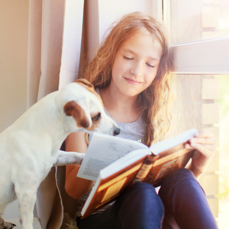 Child with dog reading book at home. Girl with pet sitting at window at readの写真素材