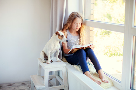 Child reading book at home. Girl sitting at window at readの写真素材