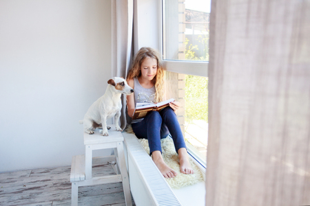 Child reading book at home. Girl sitting at window at readの写真素材