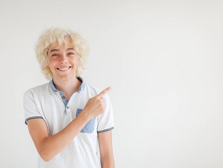 Portrait of cheerful young blond man smiling looking at camera. Happy teen at studio pointing finger...
